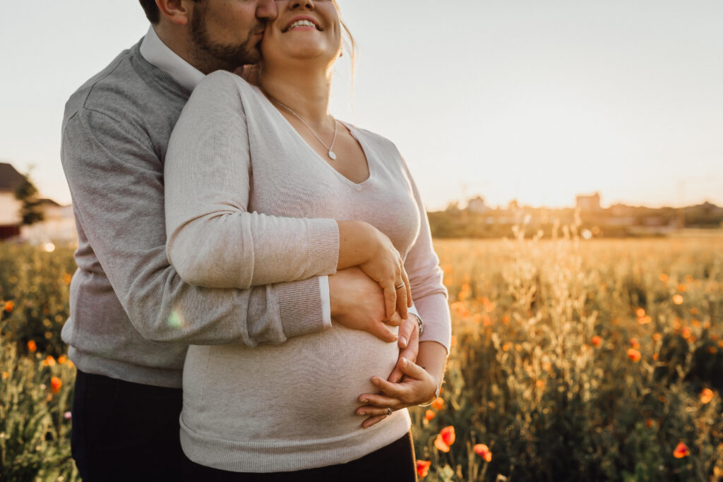 Babybauchfotos in Laatzen. Glückliches Paar mit Babybauch steht auf einem Mohnfeld und das warme Abendlicht strahlt Sommer aus