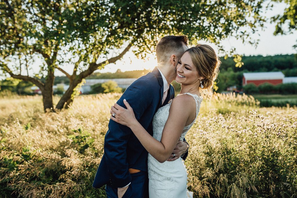 Amerikanisches Hochzeitspaar vor Wiese und Baum mit Scheune im Hintergrund. Das Abendlicht scheint und sie umarmen sich lachend.