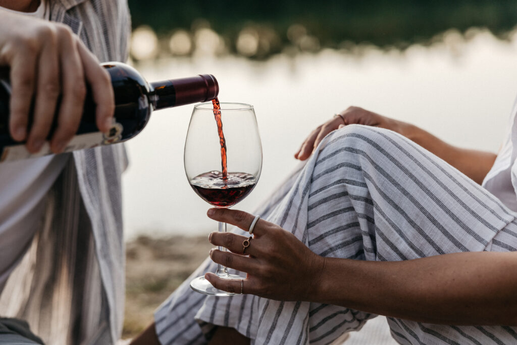 Detailaufnahme beim Einschenken von Rotwein in ein Rotweinglas, vor dem Hintergrund der Sonnenuntergangspiegelung im Wasser