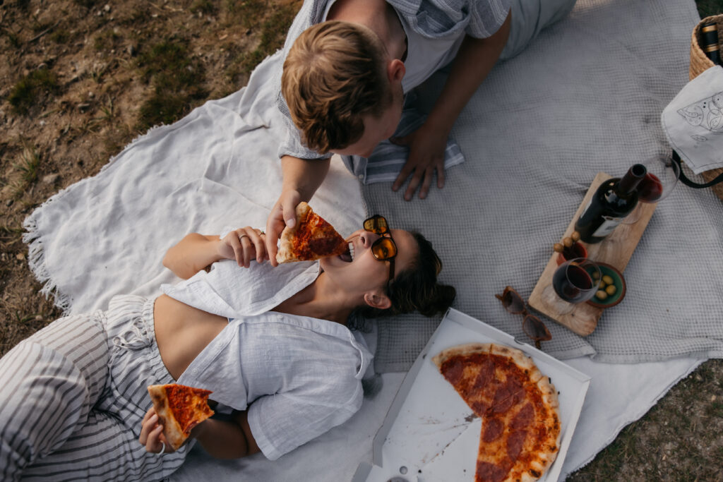 Frau liegt auf dem Rücken auf einer Decke und der Mann füttert sie mit einem Stück Pizza